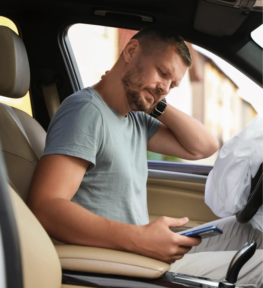 A man looking frustrated at his phone while sitting in the drivers seat of a car where the steering wheel airbag went off.