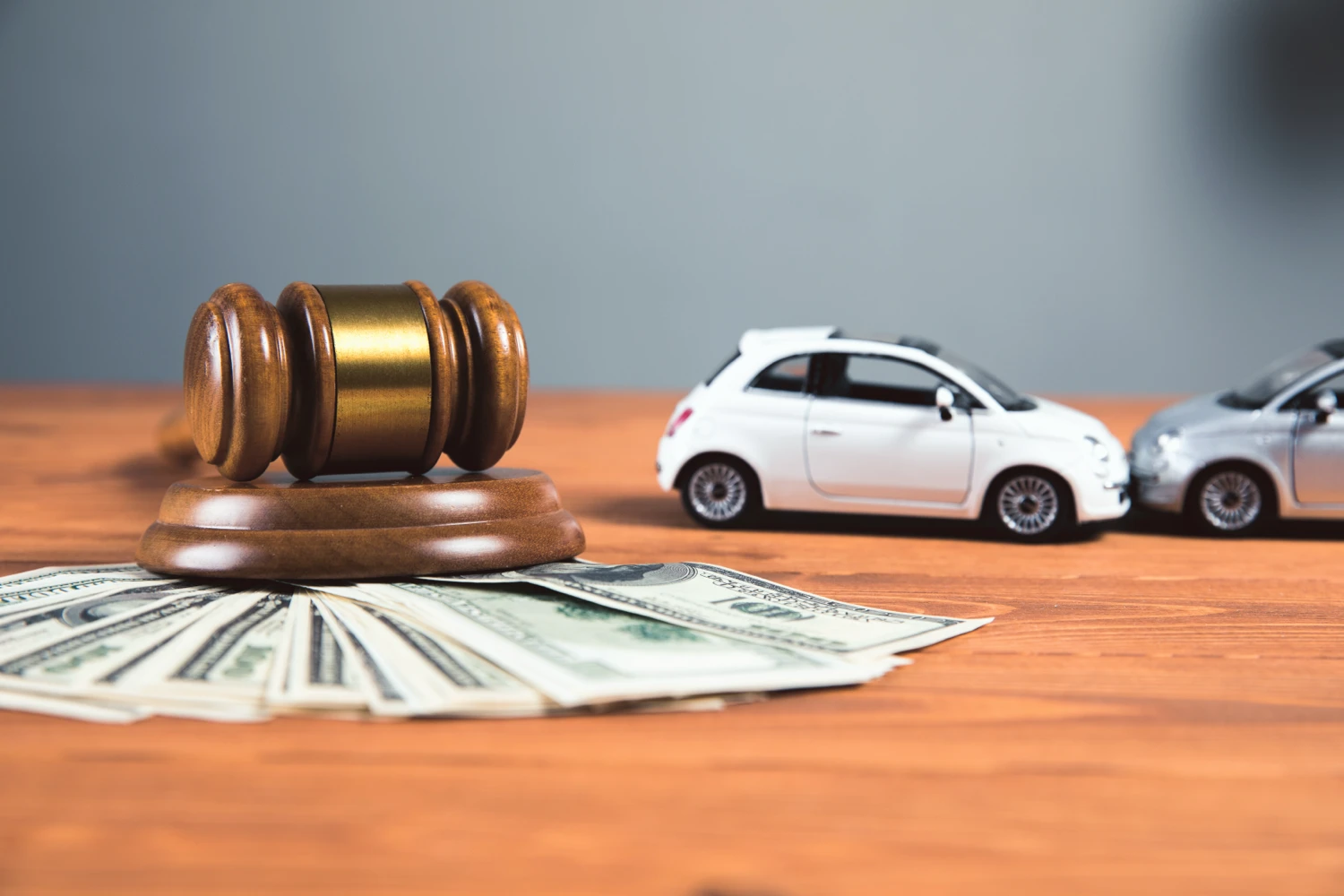 A desk with a gavel, a stack of cash, and two toy cars who look like they are in a head-on collision.